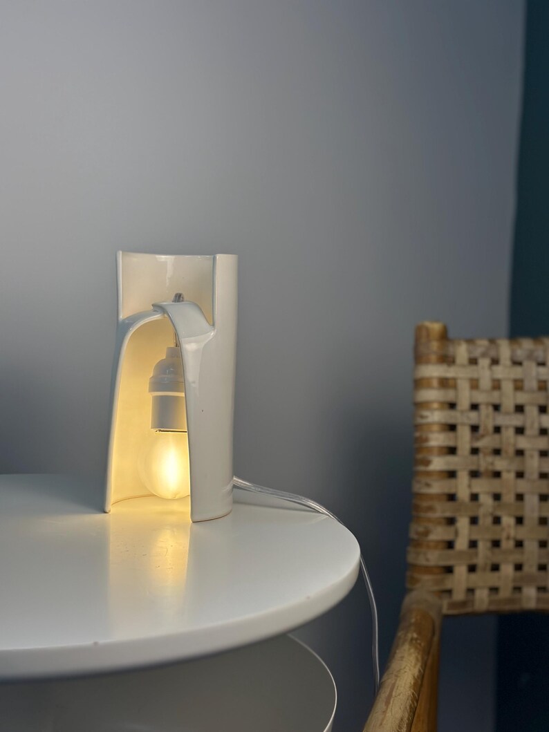 May include: A white ceramic table lamp with a curved, cylindrical structure that features an open front. The lamp houses a single light bulb suspended from the top interior, emitting a warm glow through the opening of the white ceramic body.