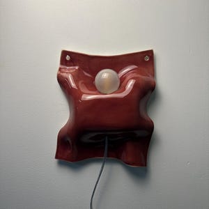 K&ouml;nnte beinhalten: Eine Wandleuchte mit einem gl&auml;nzenden, dunkelroten Keramikschirm in flie&szlig;ender, stoffartiger Form. Der Schirm verf&uuml;gt &uuml;ber zwei kleine L&ouml;cher an den oberen Ecken zur Befestigung und eine zentrale &Ouml;ffnung mit einer milchigen Glaskugel.