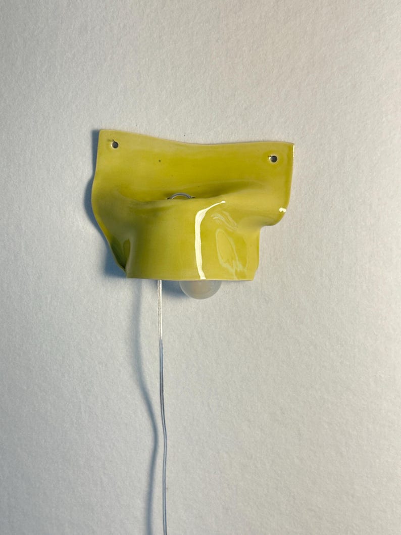 May include: A wall-mounted, chartreuse-colored ceramic lamp with a glossy finish. The lamp has a unique, organic shape with a small light bulb hanging below. A silver-colored wire extends downward from the lamp.
