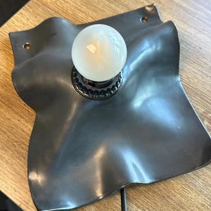 May include: A wall-mounted lamp featuring a matte black, square-shaped metal base with undulating, wavy edges. The base has two circular mounting holes at opposite corners. A single, round, frosted white light bulb is centered in the fixture.