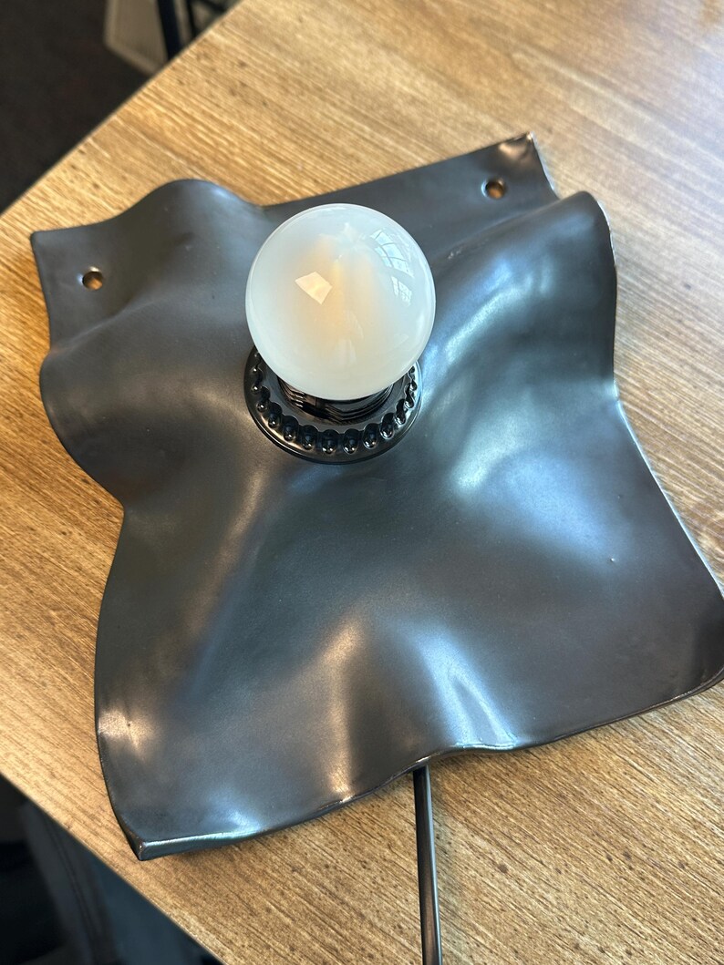 May include: A wall-mounted lamp featuring a matte black, square-shaped metal base with undulating, wavy edges. The base has two circular mounting holes at opposite corners. A single, round, frosted white light bulb is centered in the fixture.
