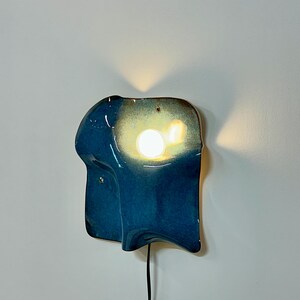 May include: A wall-mounted ceramic lamp with an irregular, organic shape and a glossy deep blue glaze. The fixture features a central circular cutout that emits a warm, bright light, casting a soft glow against the wall behind the structure.