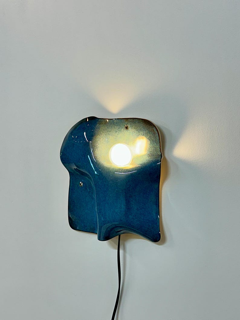 May include: A wall-mounted ceramic lamp with an irregular, organic shape and a glossy deep blue glaze. The fixture features a central circular cutout that emits a warm, bright light, casting a soft glow against the wall behind the structure.