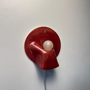 May include: A wall-mounted lamp featuring a glossy, deep red, circular ceramic base with an organic, curved protrusion. A single, spherical, frosted white light bulb is positioned within the centre of the form, connected by a thin, light-coloured cord.