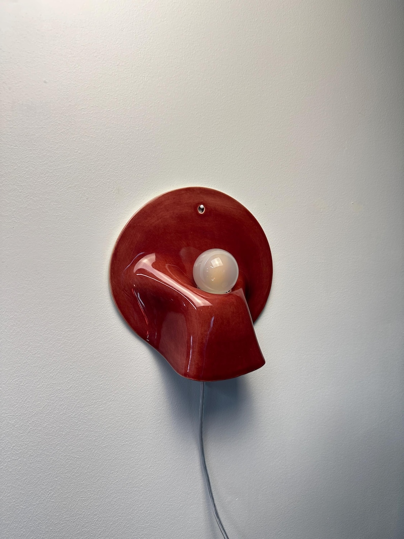 May include: A wall-mounted lamp featuring a glossy, deep red, circular ceramic base with an organic, curved protrusion. A single, spherical, frosted white light bulb is positioned within the centre of the form, connected by a thin, light-coloured cord.