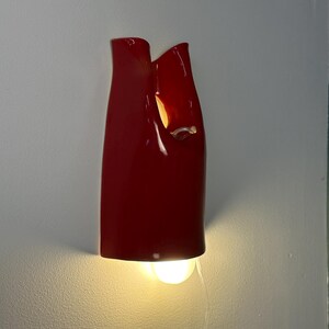 May include: A glossy red wall sconce with a smooth, curved, vertical form featuring an open top and a cutout section. The fixture emits a warm, glowing light from a bulb positioned at the bottom, casting a soft illumination downward.