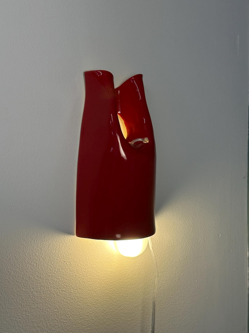 May include: A glossy red wall sconce with a smooth, curved, vertical form featuring an open top and a cutout section. The fixture emits a warm, glowing light from a bulb positioned at the bottom, casting a soft illumination downward.