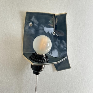 May include: A ceramic wall sconce with a dark teal glaze. The sconce features a curved shelf holding a round, frosted light bulb. The bulb is set in a black socket, and a silver cord extends downward. The sconce is mounted on a light-colored wall.
