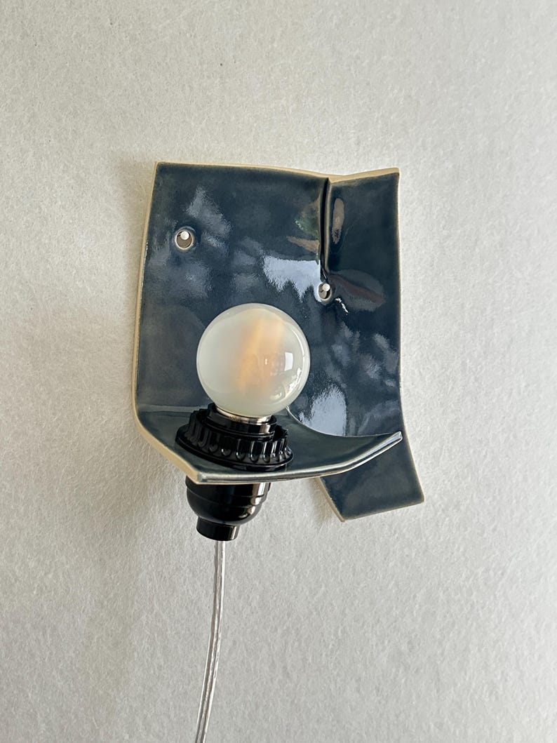 May include: A ceramic wall sconce with a dark teal glaze. The sconce features a curved shelf holding a round, frosted light bulb. The bulb is set in a black socket, and a silver cord extends downward. The sconce is mounted on a light-colored wall.