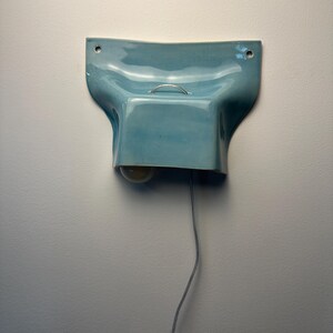 K&ouml;nnte beinhalten: Eine hellblaue Wandleuchte aus Keramik mit gl&auml;nzender Oberfl&auml;che, die eine geschwungene, geformte Gestalt mit zwei Befestigungsl&ouml;chern an den oberen Ecken aufweist. Ein kleiner silberner Metallgriff ist mittig auf der Oberseite angebracht.