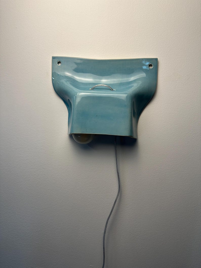 K&ouml;nnte beinhalten: Eine hellblaue Wandleuchte aus Keramik mit gl&auml;nzender Oberfl&auml;che, die eine geschwungene, geformte Gestalt mit zwei Befestigungsl&ouml;chern an den oberen Ecken aufweist. Ein kleiner silberner Metallgriff ist mittig auf der Oberseite angebracht.