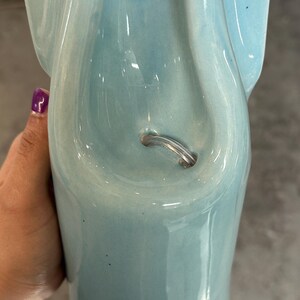 May include: A light blue ceramic wall pocket with a glossy finish. The object features a smooth, curved design with a circular hole at the top for mounting and a small, arched metal wire loop integrated into the center for hanging or display purposes.