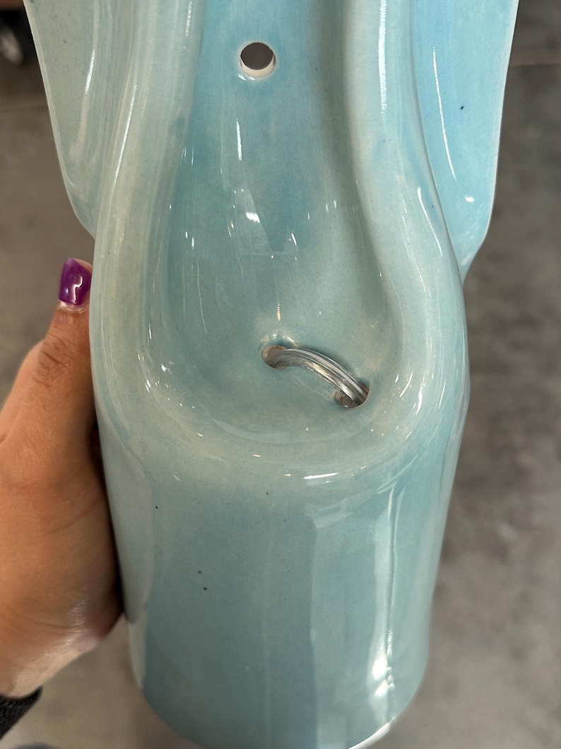 May include: A light blue ceramic wall pocket with a glossy finish. The object features a smooth, curved design with a circular hole at the top for mounting and a small, arched metal wire loop integrated into the center for hanging or display purposes.