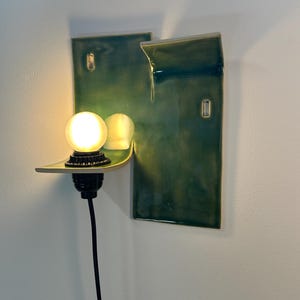 May include: A wall-mounted ceramic sconce featuring a glossy, deep green glaze with a rectangular backplate and a protruding shelf. A black socket holds a glowing, round white light bulb, and a black power cord extends downward from the fixture.