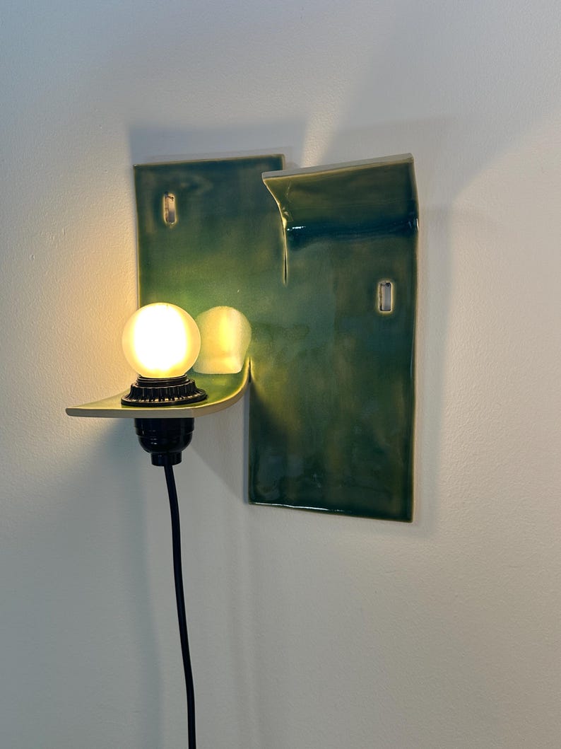 May include: A wall-mounted ceramic sconce featuring a glossy, deep green glaze with a rectangular backplate and a protruding shelf. A black socket holds a glowing, round white light bulb, and a black power cord extends downward from the fixture.