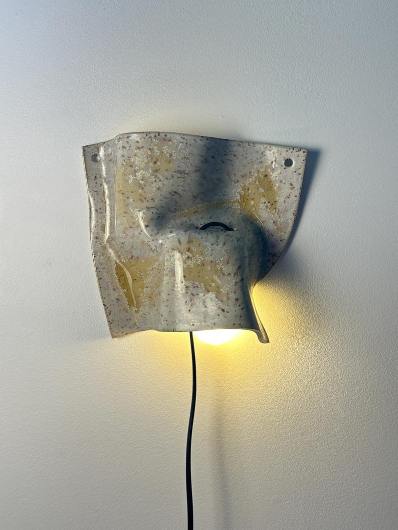 May include: A wall-mounted ceramic light fixture with a speckled off-white and tan glaze. The piece has an irregular, folded, sheet-like shape with two small holes at the top corners and a central opening emitting a warm glow from a light bulb.