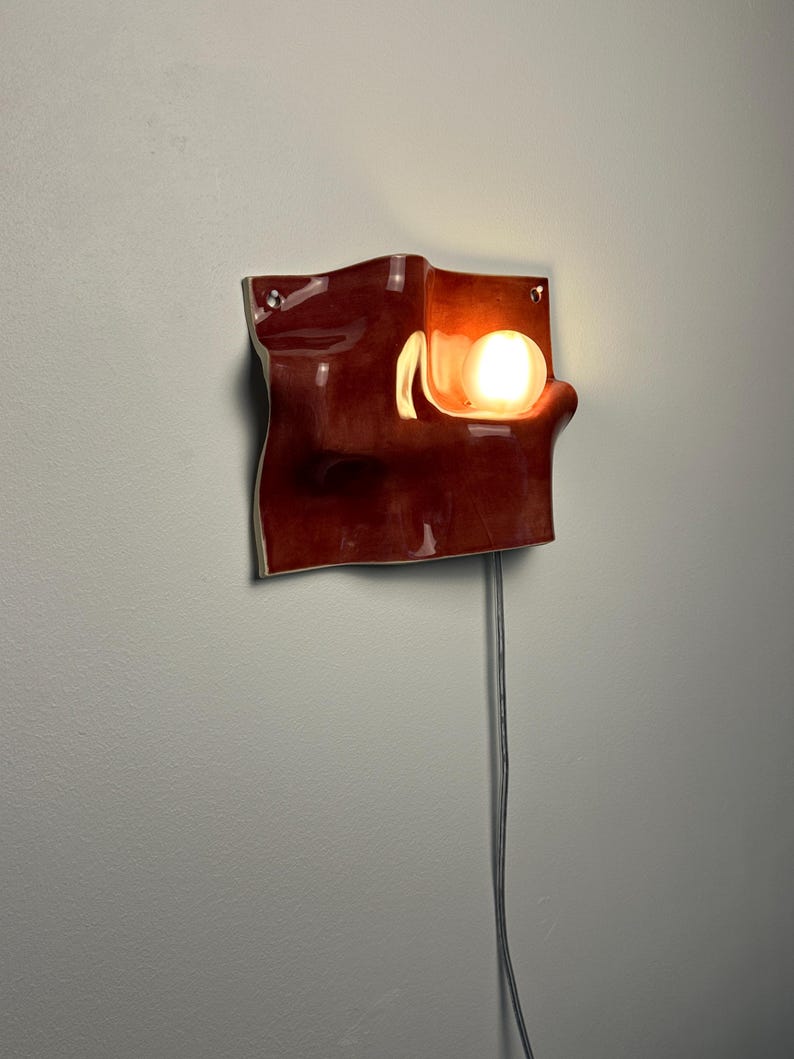 May include: A wall-mounted lamp featuring a glossy, deep red ceramic shade with an undulating, wave-like surface. A single round, glowing light bulb is recessed into a curved section of the ceramic, and a thin black power cord hangs downward.