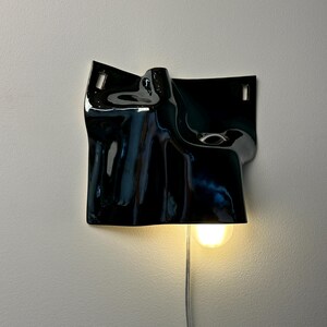 May include: A wall-mounted lamp featuring a glossy black ceramic shade with an irregular, draped, and folded surface. The fixture includes two rectangular mounting slots at the top and a single illuminated light bulb emitting a warm glow.