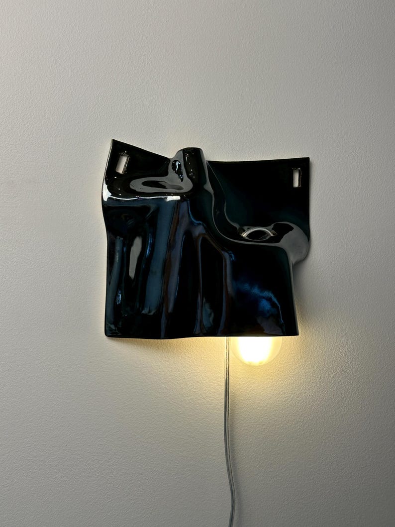 May include: A wall-mounted lamp featuring a glossy black ceramic shade with an irregular, draped, and folded surface. The fixture includes two rectangular mounting slots at the top and a single illuminated light bulb emitting a warm glow.