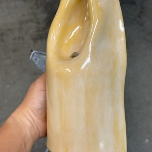 Pu&ograve; includere: Una base per lampada in ceramica alta e verticale con una finitura lucida color giallo pallido e crema. La struttura presenta una forma organica ondulata con una profonda rientranza curva nella parte superiore e un portalampada elettrico visibile.