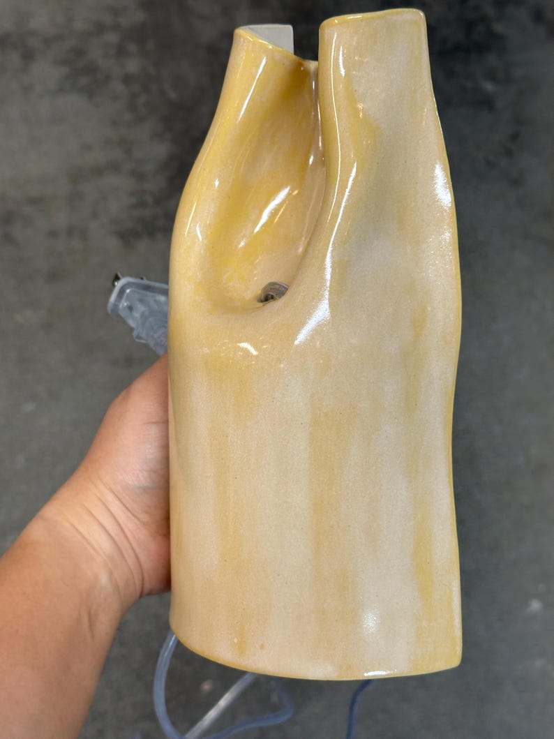 Pu&ograve; includere: Una base per lampada in ceramica alta e verticale con una finitura lucida color giallo pallido e crema. La struttura presenta una forma organica ondulata con una profonda rientranza curva nella parte superiore e un portalampada elettrico visibile.