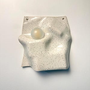 May include: A wall-mounted ceramic light fixture with an off-white, speckled glaze and an irregular, undulating surface. A single, spherical, frosted glass light bulb is embedded into the center of the textured, wavy ceramic form.