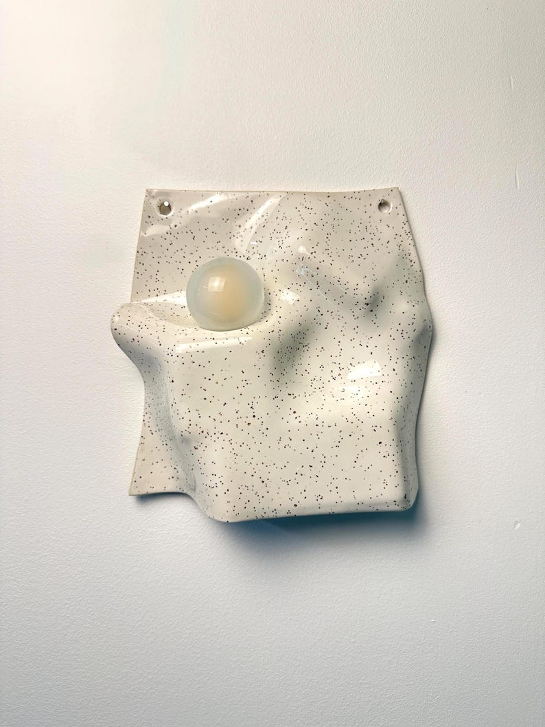 May include: A wall-mounted ceramic light fixture with an off-white, speckled glaze and an irregular, undulating surface. A single, spherical, frosted glass light bulb is embedded into the center of the textured, wavy ceramic form.
