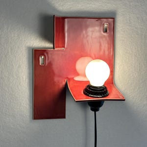 Pode incluir: Uma moderna arandela de parede vermelha com um design geom&eacute;trico &uacute;nico. A arandela tem um acabamento brilhante e uma pequena l&acirc;mpada redonda. O cabo preto pende para baixo. A arandela &eacute; montada em uma parede cinza.