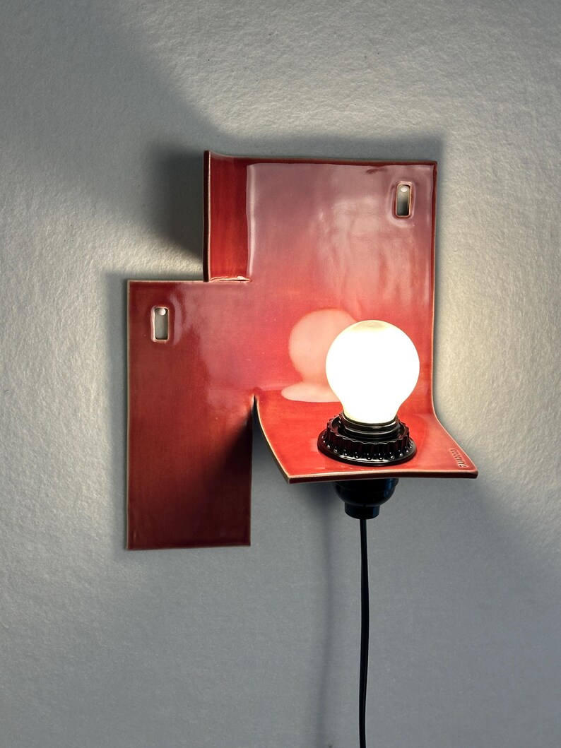 Pode incluir: Uma moderna arandela de parede vermelha com um design geom&eacute;trico &uacute;nico. A arandela tem um acabamento brilhante e uma pequena l&acirc;mpada redonda. O cabo preto pende para baixo. A arandela &eacute; montada em uma parede cinza.