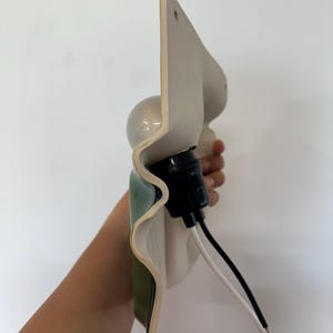 May include: A wall-mounted light fixture featuring a wavy, off-white ceramic shade with a green interior finish. The fixture includes a black socket, a round light bulb, and two exposed electrical wires, one black and one white, extending outward.