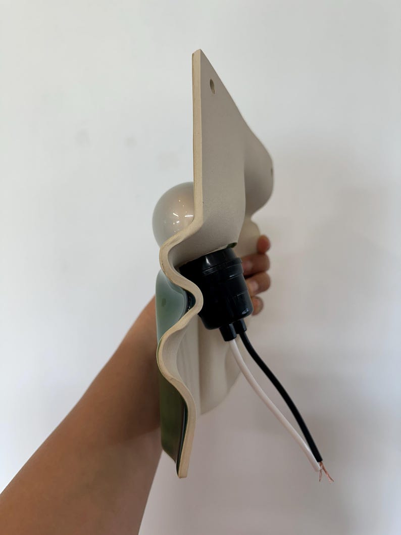 May include: A wall-mounted light fixture featuring a wavy, off-white ceramic shade with a green interior finish. The fixture includes a black socket, a round light bulb, and two exposed electrical wires, one black and one white, extending outward.