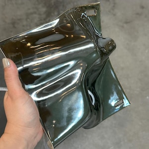 May include: A glossy, dark olive green ceramic wall sconce with a fluid, undulating surface. The rectangular piece features a prominent central protrusion and two small, rectangular mounting slots located at the top and bottom edges.