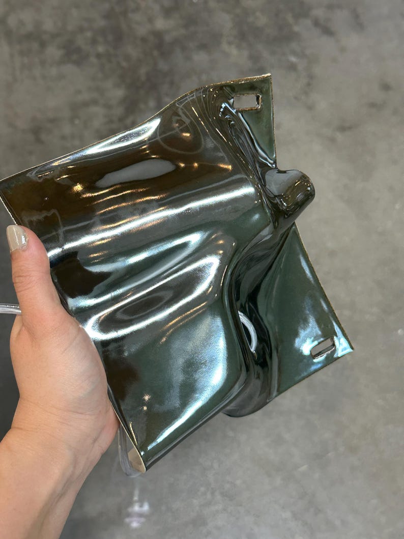 May include: A glossy, dark olive green ceramic wall sconce with a fluid, undulating surface. The rectangular piece features a prominent central protrusion and two small, rectangular mounting slots located at the top and bottom edges.