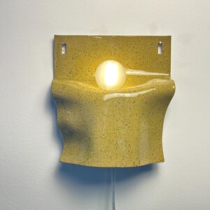 May include: A wall-mounted lamp featuring a yellow ceramic base with a speckled finish and a wavy, folded design. A single, round, frosted glass light bulb is positioned in the center of the ceramic structure, emitting a warm, soft glow.