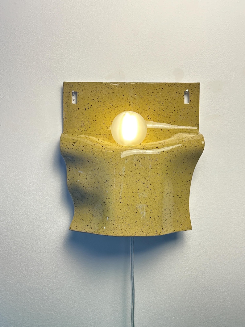May include: A wall-mounted lamp featuring a yellow ceramic base with a speckled finish and a wavy, folded design. A single, round, frosted glass light bulb is positioned in the center of the ceramic structure, emitting a warm, soft glow.