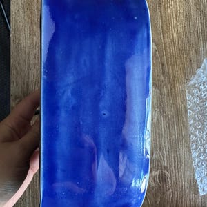 Może przedstawiać: Prostokątny, kobaltowoniebieski ceramiczny pojemnik o zaokrąglonych krawędziach. Błyszcząca powierzchnia odbija światło, podkreślając wariacje kolorystyczne. Pojemnik jest trzymany na drewnianej powierzchni.