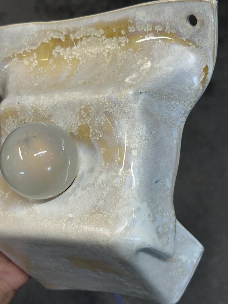 May include: A wall-mounted ceramic light fixture featuring a glossy, off-white glaze with a textured, crackled finish revealing amber-toned undertones. The fixture includes a single, smooth, spherical glass bulb protruding from the center.
