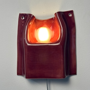 Puede incluir: Un aplique de pared de cer&aacute;mica con un esmalte brillante de color burdeos intenso. La pieza rectangular presenta una cavidad central empotrada que alberga una bombilla encendida, con dos peque&ntilde;os orificios circulares de montaje en las esquinas superiores y un cable.