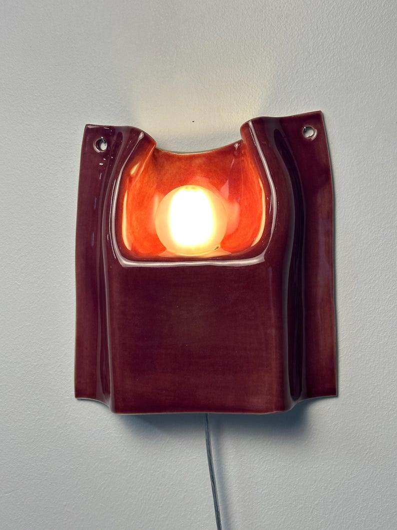 Puede incluir: Un aplique de pared de cer&aacute;mica con un esmalte brillante de color burdeos intenso. La pieza rectangular presenta una cavidad central empotrada que alberga una bombilla encendida, con dos peque&ntilde;os orificios circulares de montaje en las esquinas superiores y un cable.
