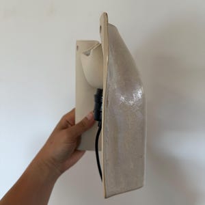 May include: A wall-mounted ceramic light fitting with a glossy, off-white finish. The structure features a curved, elongated shape with a flat back panel and a protruding socket area. A black electrical cable extends from the central light socket.