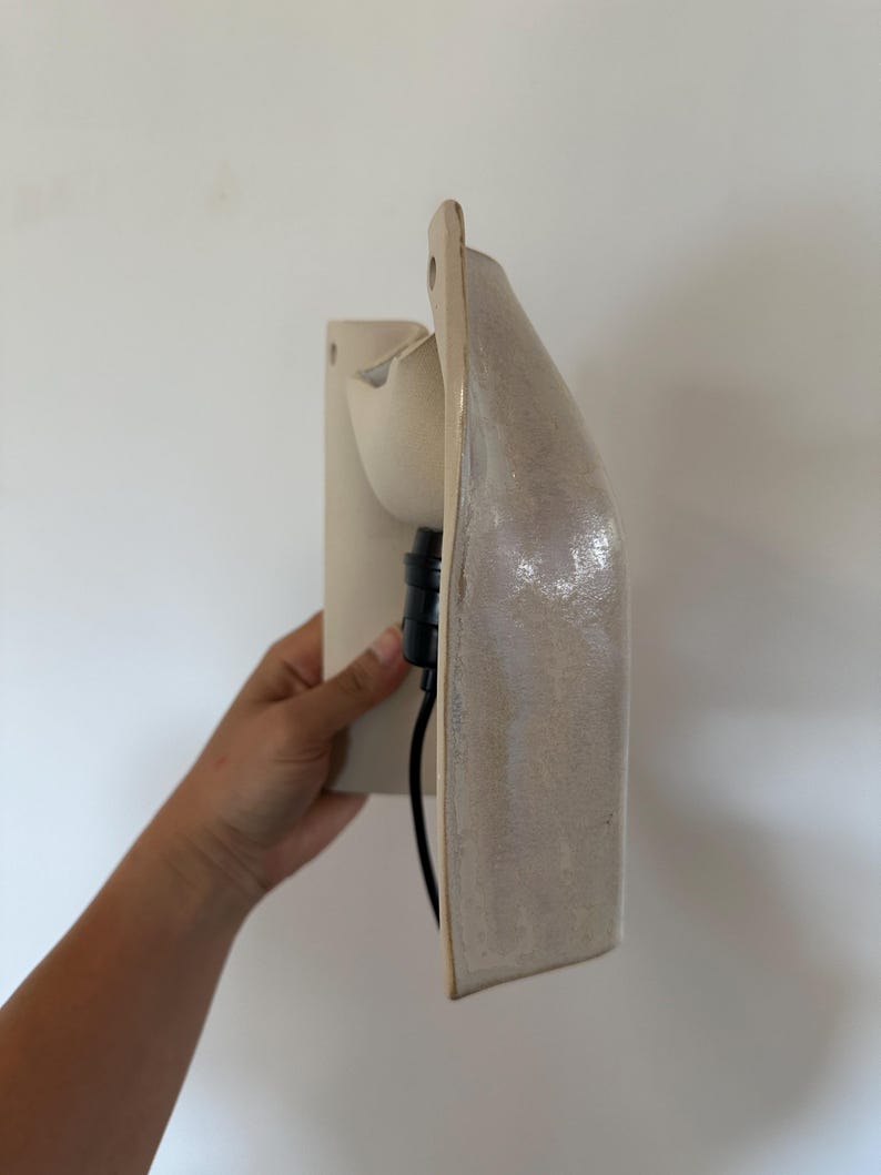 May include: A wall-mounted ceramic light fitting with a glossy, off-white finish. The structure features a curved, elongated shape with a flat back panel and a protruding socket area. A black electrical cable extends from the central light socket.