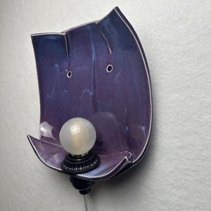 May include: A wall-mounted ceramic sconce featuring a glossy, deep purple glaze with subtle blue undertones. The curved, organic-shaped backplate holds a single black socket with a round, frosted white light bulb and a clear power cord extending below.
