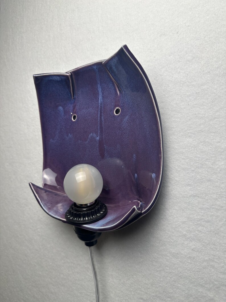 May include: A wall-mounted ceramic sconce featuring a glossy, deep purple glaze with subtle blue undertones. The curved, organic-shaped backplate holds a single black socket with a round, frosted white light bulb and a clear power cord extending below.