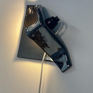 Peut inclure: Une applique murale compos&eacute;e d'une base en c&eacute;ramique au fini chrom&eacute; r&eacute;fl&eacute;chissant, pr&eacute;sentant une forme sculpturale angulaire. Le luminaire comprend une douille noire pour ampoule et un cordon d'alimentation transparent descendant du centre.