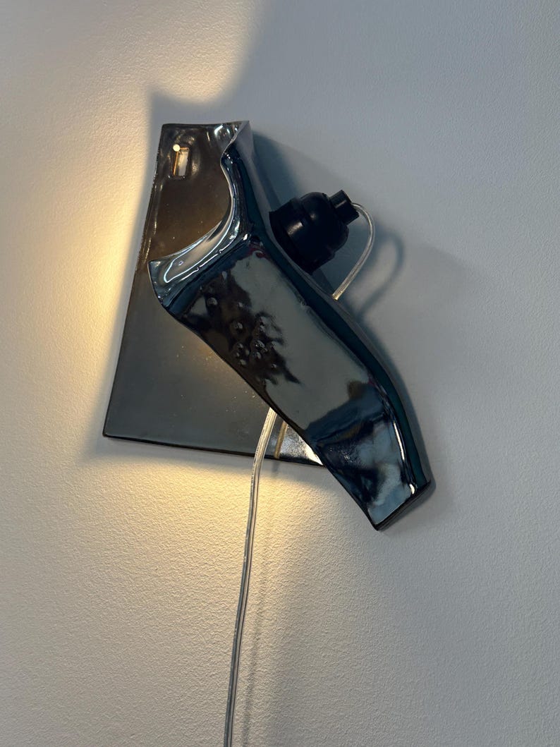 Peut inclure: Une applique murale compos&eacute;e d'une base en c&eacute;ramique au fini chrom&eacute; r&eacute;fl&eacute;chissant, pr&eacute;sentant une forme sculpturale angulaire. Le luminaire comprend une douille noire pour ampoule et un cordon d'alimentation transparent descendant du centre.