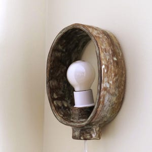 May include: A ceramic wall sconce with a brown and green mottled glaze. The sconce has an oval shape with a white light bulb in the center. A white cord hangs down from the bottom.