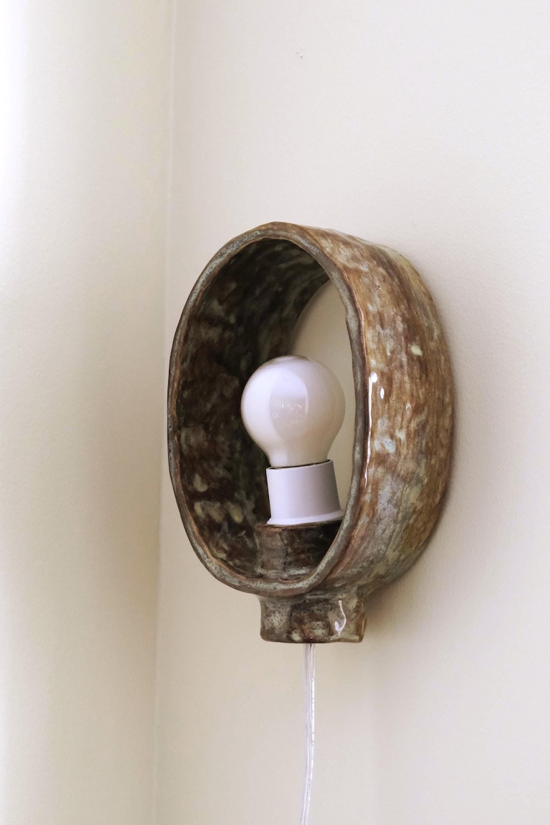 May include: A ceramic wall sconce with a brown and green mottled glaze. The sconce has an oval shape with a white light bulb in the center. A white cord hangs down from the bottom.