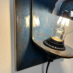 May include: A wall-mounted lamp featuring a rectangular ceramic backplate with a glossy, mottled blue and dark brown glaze. A curved shelf extends from the base, holding a black socket with a clear glass light bulb that has a silver-dipped top.