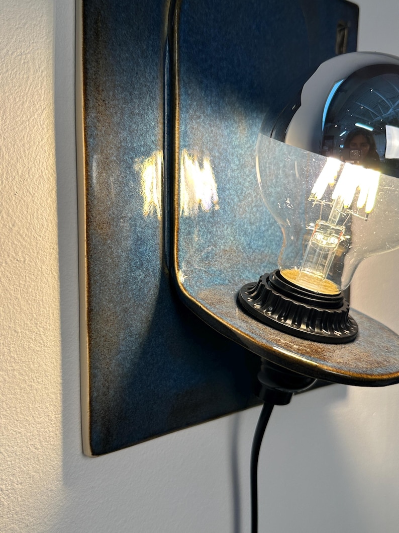 May include: A wall-mounted lamp featuring a rectangular ceramic backplate with a glossy, mottled blue and dark brown glaze. A curved shelf extends from the base, holding a black socket with a clear glass light bulb that has a silver-dipped top.