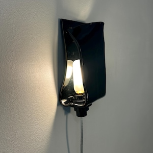 May include: A wall-mounted lamp featuring a glossy black ceramic shade with a curved, folded design. The fixture holds a single glowing light bulb in the center, casting a warm light against the wall, and includes a clear power cord extending downward.